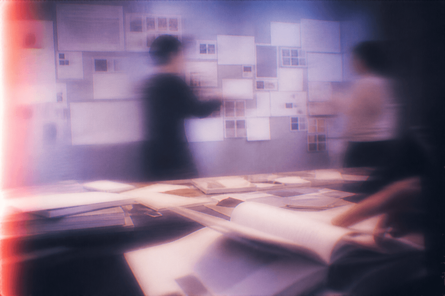 Blurred figures beside a paper-covered table and a wall filled with overlapping sheets, lit in purple and red
